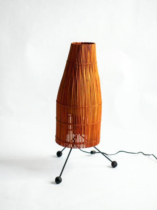 Tapered woven rattan fish trap table lamp on a dark metal base with splayed legs and round feet.